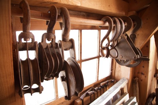 Hanging rusted heavy cast iron hooks attached to pulley wheel on wood rod by window light in traditional rustic industrial woodworking workshop