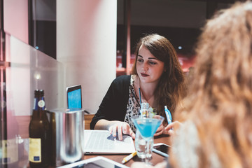 Two young beautiful caucasian women sitting in a bar using computer and smart phone hand hold, discussing - technology, business, studying concept