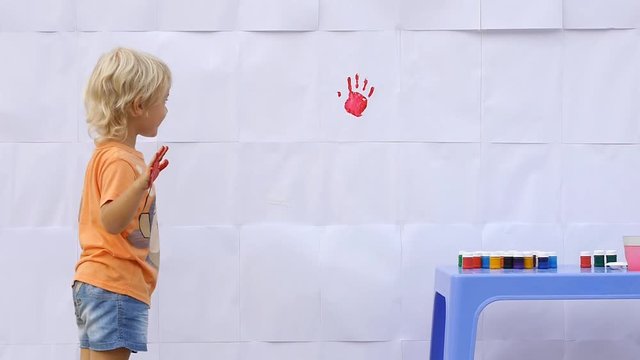 Happy little cute child painting first color handprints on the white wall. Showing his palm and waving hands in colorful print 50fps
