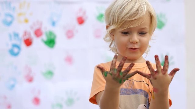 Amazing cute happy child surprising, saying wow and rubbing color paint on his little hands on color handprints background 50fps
