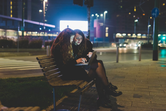 Two young beautiful caucasian women friends outdoor in the city night using computer, tapping keyboard - business, technology, social network concept