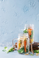 Variety of vegetable salads in mason jars. Salad with greens, pasta, carrots, cauliflower, salmon. Standing with wooden spoon and serving board over blue texture background. Healthy food to go.