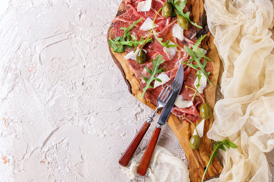 Beef carpaccio on olive wood serving board with capers, olive oil cheese and arugula, served with knife, fork, gauze textile over beige concrete texture background. Top view with space for text.