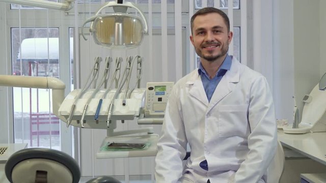 Brunette caucasian dentist sitting down on the chair at his office. Middle aged male doctor smiling for the camera against background of dental equipment. Handsome bearded man in white coat and