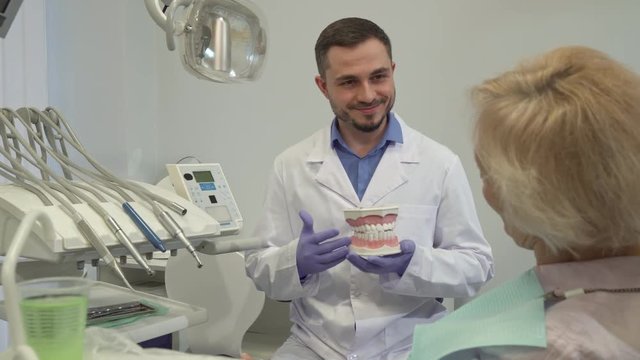 Brunette caucasian dentist explaining something on layout of teeth to his female client. Senior blond woman sitting on dental chair backwards to the camera. Handsome male doctor standing near the