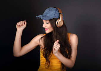 Beautiful emotional dancing and singing young woman listening the music in wireless headphone in blue cap and yellow top on dark grey background. Closeup portrait