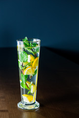 Glass of lemonade with mint and lemon wedges on the dark wooden background. Selective focus