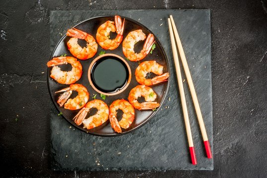 Seafood. Shellfish. Fried grilled shrimp prawns on wooden skewers, with soy sauce, asian style. Snack, appetizer. On black concrete table, copy space top view