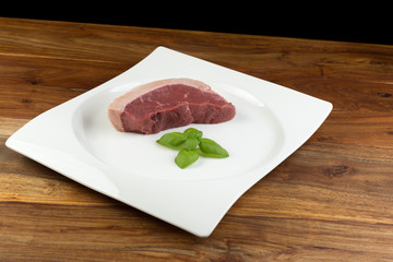 Slice of raw cow beef (picanha) and basil leaves on a white dish bow and natural wood table - black background
