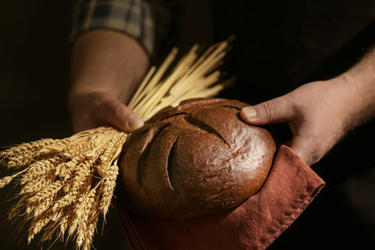 Male hands with loaf of bread and spikelets on dark background, closeup