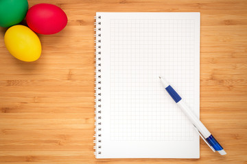 Red, green, yellow Easter eggs on a light wooden background. Eggs in the upper left corner. The concept of a holiday and a happy Easter. Top view, pen notepad in the center with place for text