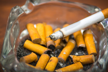 Cigarettes and tobacco lying inside and around glass ash tray on wooden surface, seen from above, anti smoking concept