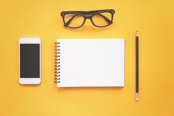 Flat lay design of work desk with notebook, glasses, pencil and smartphone on yellow background.