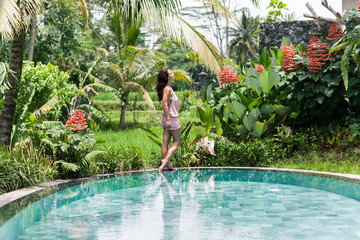 Fashion photo of beautiful tanned woman with brown hair in elegant pink dress relaxing beside a swimming pool