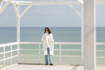 Young woman (brunette) in a white coat on the promenade near the sea.