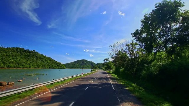 Camera Moves on Road along River by Hilly Landscape past Houses