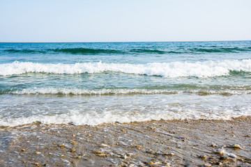 Sea tidal waves with white foam on a sunny sandy beach in resort on summer vacation. Background of ocean water texture with brown pebble (small stones) in the foreground