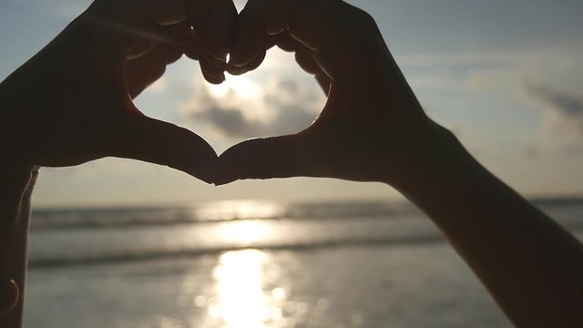 Girl making heart with her hands over sea background with beautiful golden sunset. Silhouette of female arm in heart shape with sunrise inside. Vacation concept. Summer holidays on beach. Slow motion