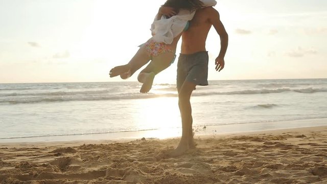 Young couple on the beach, man hug and spin around his woman on sunset. Girl jumps into her boyfriend arms, he whirling her at beautiful seaside. Having fun together at vacation. Slow motion