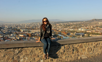Fototapeta premium Traveler girl wearing black leather jacket sitting on the stone wall. Panoramic view of old Tbilisi, Georgia at the background. The view from the castle Narikala.