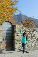 Fototapeta premium Traveler girl with hands up walking near old stone wall. Jvari Monastery on the hill at the background. Woman traveler walking in city Mtskheta, Georgia