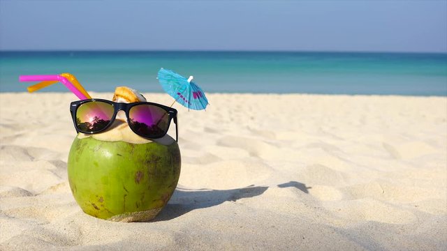 Fresh coconut with drinking straw and sunglasses on sandy white beach. With empty space for text. Dolly shot UHD