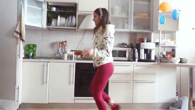 Joyful young beautiful woman is dancing in kitchen wearing pajamas and headphones drinks a cup of coffee in the morning listening to music on smartphone. 3840x2160