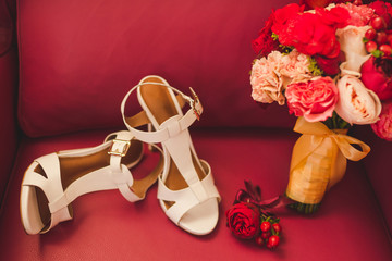 Bridal morning details. Wedding bouquet of red and pink flowers, white leather shoes and groom's boutonniere on red leather armchair.
