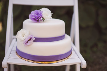 White wedding cake with purple flowers decoration outdoors on wooden modern chair. Front view, copy space.