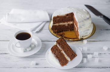 Piece of homemade cake and coffee on a white wooden table. Biscuit cake and knife at the background. Lump sugar scattered on a table.