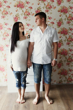 Man and pregnant woman in white shirts and jeans stand in bright room