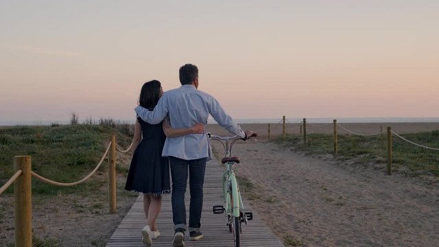 Unrecognizable couple strolls at the boardwalk on the empty beach during the soft and pink sunset, walk their mint bike, hug and cuddle each other and look into the distance
