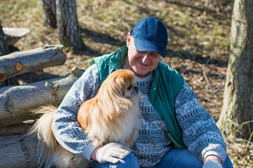 A man works in the garden, fires a bonfire, removes, cleans the territory, cuts dry branches with his little red dog near blue river