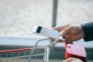 Cellphone and cart on store goods background. Closeup on person holding mobile smart phone in hand during shopping.