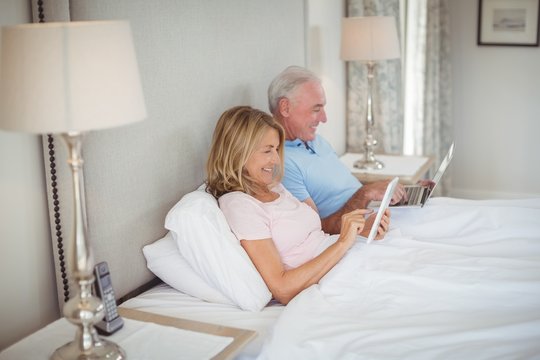 Happy senior couple on bed using laptop and digital tablet