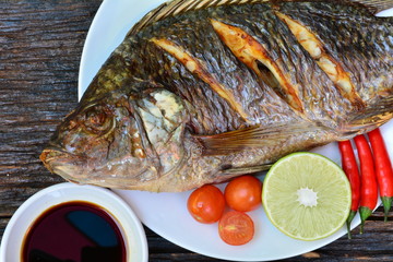 Grilled fish on white plate with chili pepper, lemon and tomato on wooden background, top view.