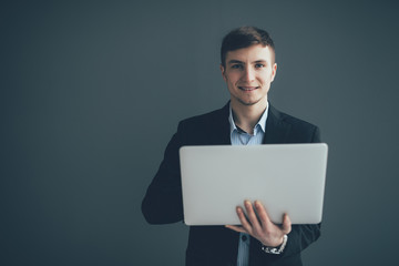 Handsome young businessman is using a laptop, looking at camera and smiling, on black background