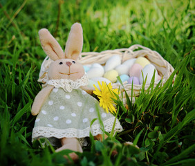 Toy bunny near a basket with egg-shaped sweets on green grass, natural light