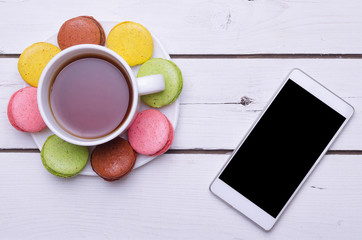 A cup of hot tea, colorful almond cookies and a smartphone on a wooden table.
