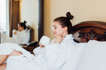 Young woman in a white bathrobe drinking tea or coffee on the bed in the hotel room