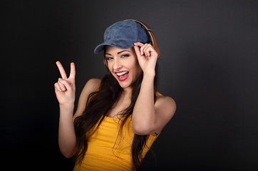 Joyful teenager listening the music in gold wireless headphone and showing victory v-sign gesture in yellow top and blue cap on dark grey background.