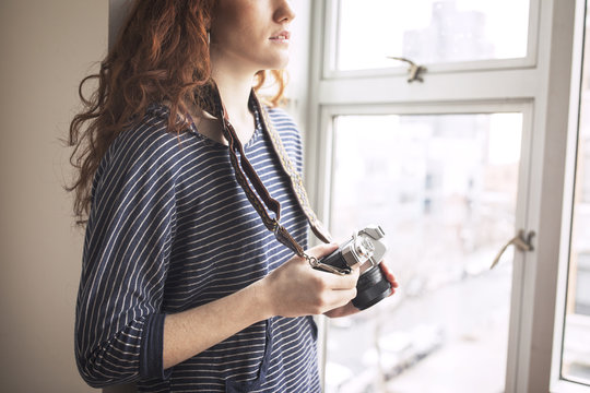 Midsection of woman with camera standing by window at home