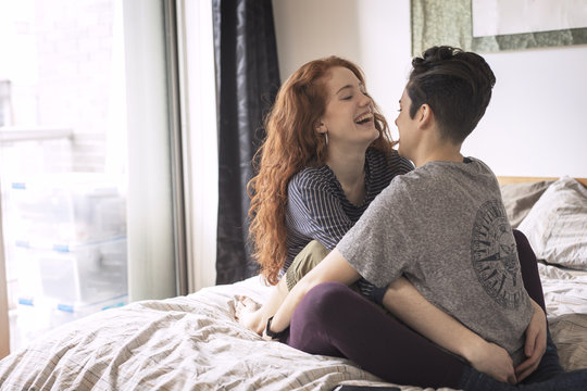 Cheerful lesbians sitting face to face on bed at home