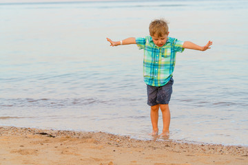 Little boy walks barefoot in shallow sea water. Tries not to wet his shorts. Evening light.