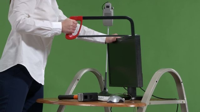 Frustrated woman office worker damaging computer with hacksaw against green screen