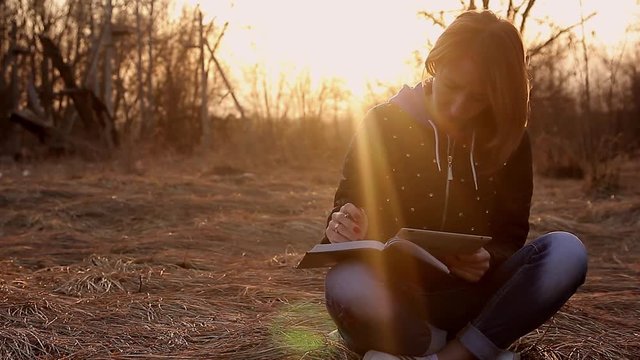 Beautiful young girl, sitting on a fallen autumn leaves in a park, reading tablet computer to take notes in a notebook, sunset light. Concept girl freelancer, working in a spring park.