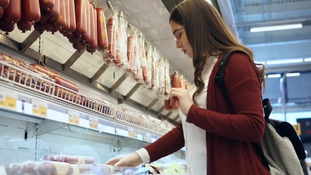 Young woman chooses sausages in grocery department in the supermarket. She carefully examines the composition of each of type, considering the label.