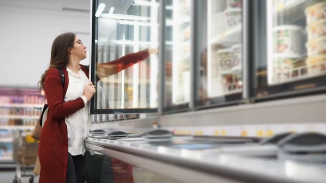 Woman with a backpack chooses products from the refrigerator in the supermarket. She carefully studies the composition and reads the label.