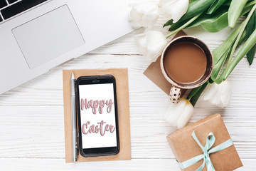 happy easter text greeting card sign on empty phone screen and laptop with morning coffee and tulips on white wooden rustic background. stylish flat lay with flowers and  gadgets