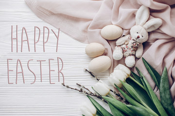happy easter text greeting card sign on stylish easter eggs and willow buds and rabbit and white tulips on rustic wooden background and fabric flat lay.  concept with space for text, top view